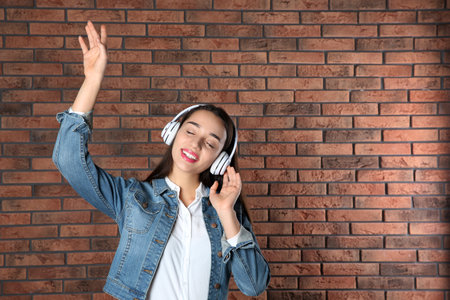 Beautiful young woman listening to music with headphones against brick wall. Space for textの写真素材