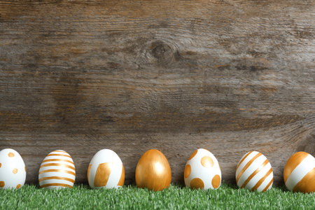 Line of traditional Easter eggs decorated with golden paint on green lawn near wooden wall, space...