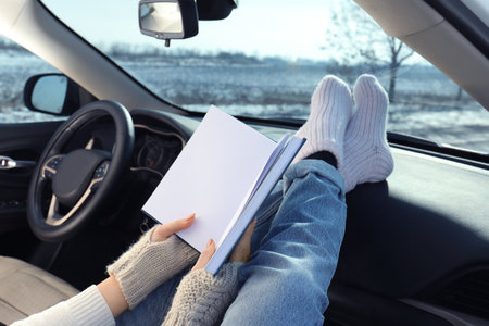 Young woman in warm socks holding her legs on car dashboard and reading book, closeup. Cozy atmosphereの写真素材