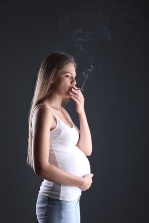 Young pregnant woman smoking cigarette on dark background. Harm to unborn babyの写真素材