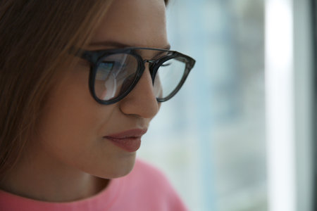 Young woman wearing glasses on blurred background, closeup. Ophthalmology service