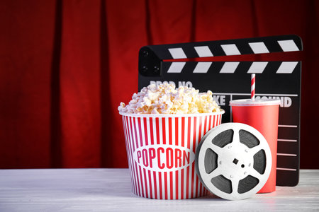 Clapperboard, reel and popcorn bucket on table against stage curtain, space for text. Watching cinemaの写真素材