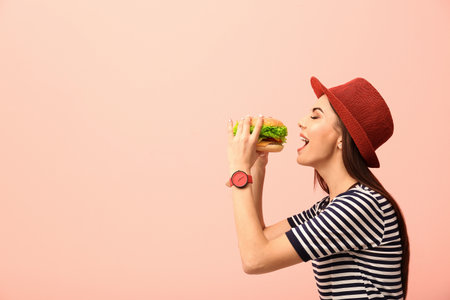 Young woman eating tasty burger on color background. Space for textの写真素材