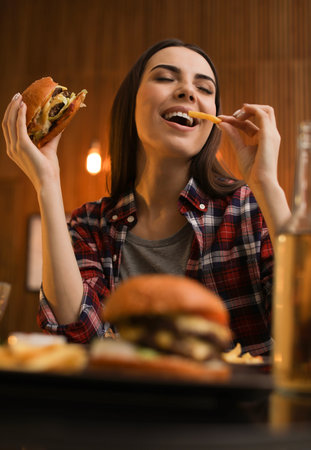 Young woman eating French fries and tasty burger in cafeの写真素材