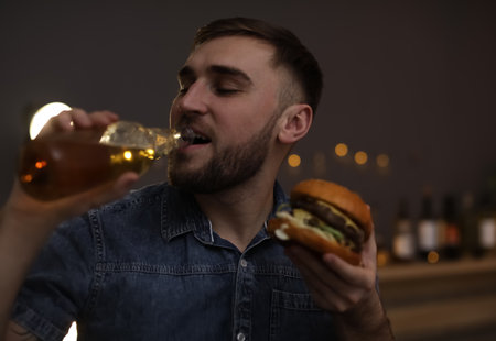 Young man with beer eating tasty burger in cafeの写真素材