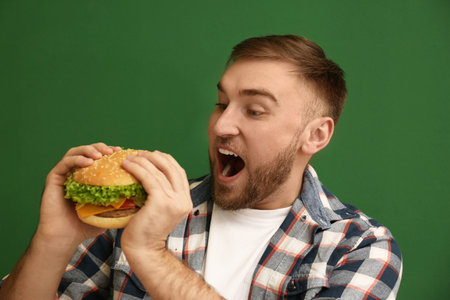 Young man eating tasty burger on color backgroundの写真素材