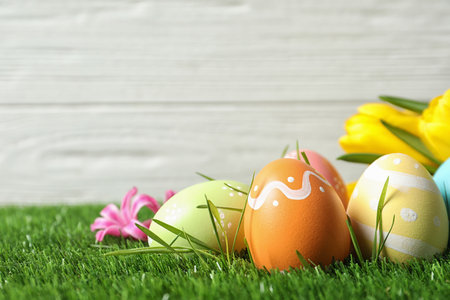 Colorful painted Easter eggs and flowers on green grass against wooden background, space for text