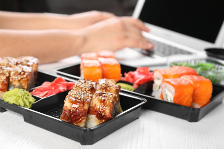 Boxes with different sushi rolls and blurred woman using laptop on background. food deliveryの写真素材