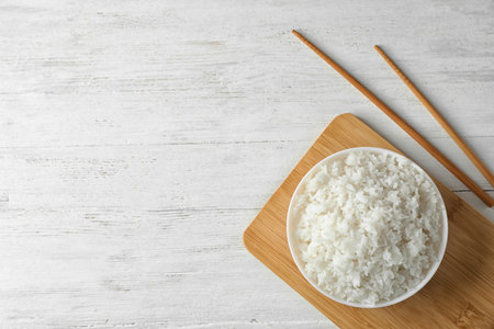 Board with boiled rice in bowl and chopsticks on wooden background, top view. Space for textの写真素材