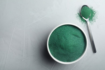 Bowl and spoon of spirulina algae powder on gray background, top view with space for text