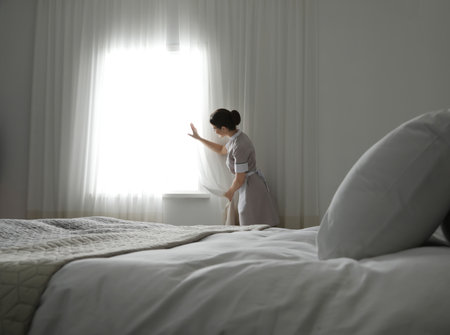Young maid opening curtains in hotel roomの写真素材