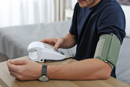 Man checking blood pressure with sphygmomanometer at table indoors, closeup. Cardiology concept
