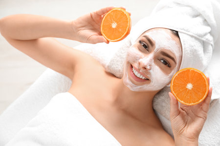 Pretty woman with rejuvenating facial mask holding sliced orange in spa salon, above viewの写真素材