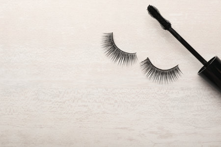 Mascara brush and artificial eyelashes on light background, top view with space for text