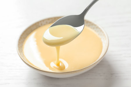 Spoon with pouring condensed milk over bowl on white background, closeup. Dairy productsの写真素材