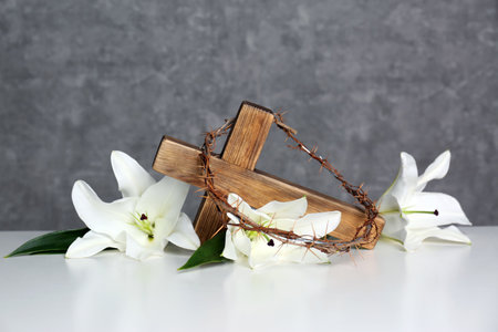 Wooden cross, crown of thorns and blossom lilies on table against color background