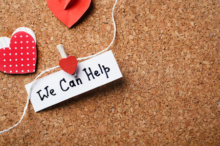 Paper hearts and note with words WE CAN HELP attached to string on cork board. Space for textの写真素材