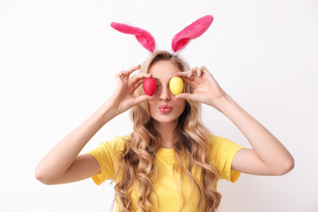 Beautiful young woman in bunny ears headband holding Easter eggs near eyes on light backgroundの写真素材