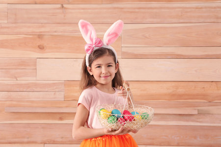 Little girl in bunny ears headband holding basket with Easter eggs against wooden background, space for textの写真素材