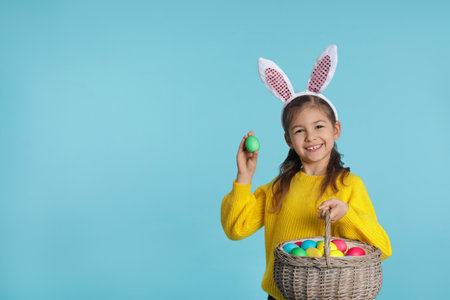 Little girl in bunny ears headband holding basket with Easter eggs on color background, space for...
