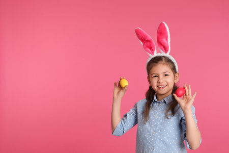 Little girl in bunny ears headband holding Easter eggs on color background, space for text