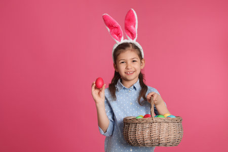 Little girl in bunny ears headband holding basket with Easter eggs on color background, space for...