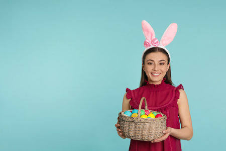 Beautiful woman in bunny ears headband holding basket with Easter eggs on color background, space for textの写真素材