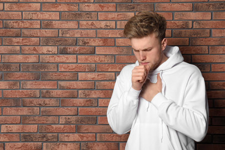 Handsome young man coughing near brick wall. Space for textの写真素材