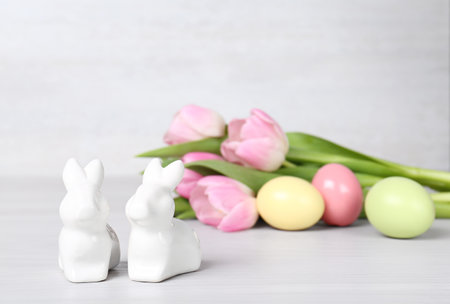 Cute ceramic bunnies, Easter eggs and spring tulips on table against light background, space for textの写真素材