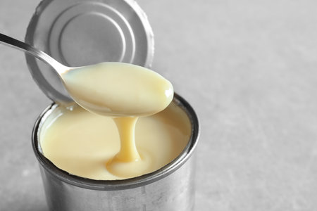 Condensed milk pouring from spoon into tin can on gray background, space for text. Dairy productの写真素材