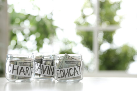 Glass jars with money for different needs on table against blurred background, space for textの写真素材