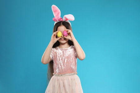 Little girl in bunny ears headband holding Easter eggs near eyes on color backgroundの写真素材