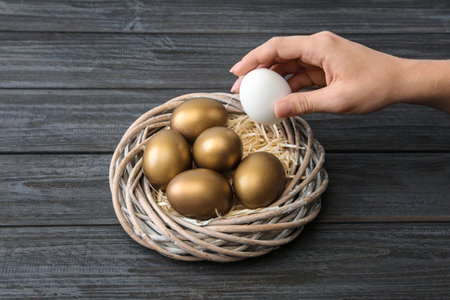 Woman taking egg from nest with gold ones on wooden tableの写真素材
