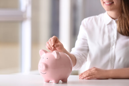 Woman putting coin into piggy bank at table indoors, closeup. Space for textの写真素材