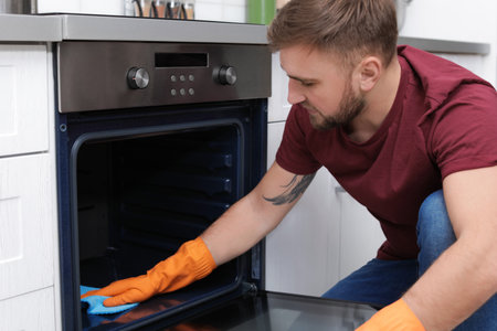 Young man cleaning oven with rag in kitchenの写真素材