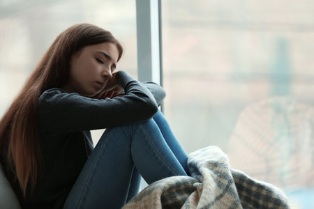 Upset teenage girl sitting at window indoors. Space for textの写真素材