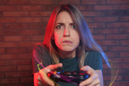 Emotional young woman playing video games with controller near brick wallの写真素材