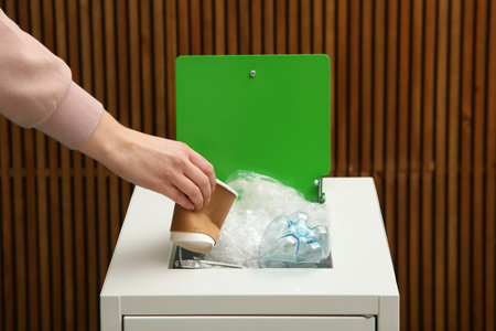 Woman putting used paper cup into trash bin on wooden background, closeup. Recycling conceptの写真素材