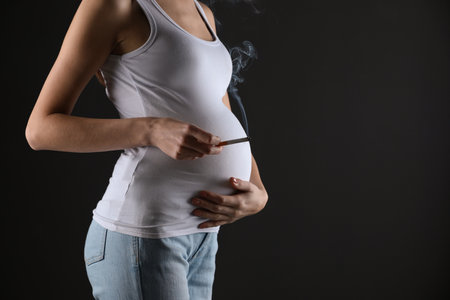 Pregnant woman smoking cigarette on black background, closeup. Space for textの写真素材