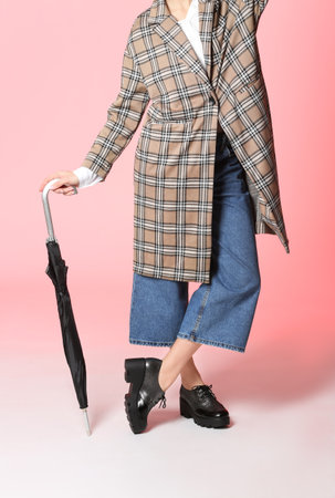 Woman in stylish shoes and coat holding umbrella on color backgroundの写真素材