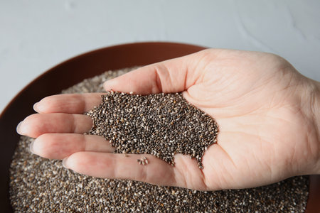 Woman holding heap of chia seeds over bowl on gray background, closeup