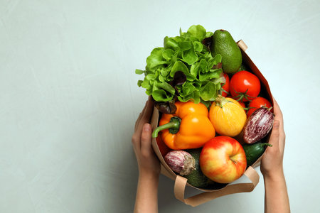 Woman holding paper bag with fresh vegetables and fruits on light background, top view. Space for textの写真素材