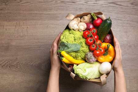 Woman holding paper package full of fresh vegetables on wooden background, top view. Space for textの写真素材