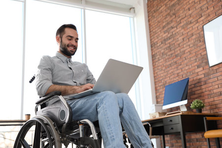 Young man in wheelchair using laptop at officeの写真素材