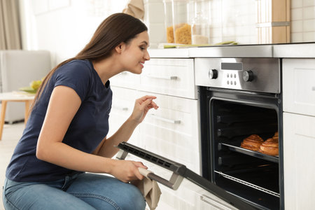 Beautiful woman opening door of oven with baked buns in kitchenの写真素材