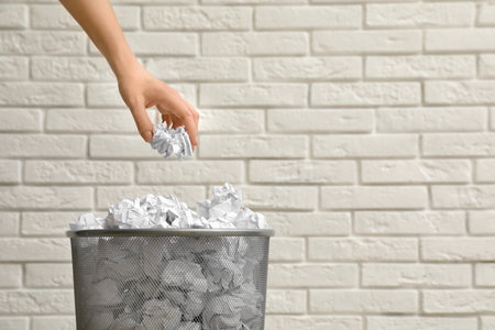 Woman throwing crumpled paper into metal bin against brick wall, closeup. Space for textの写真素材