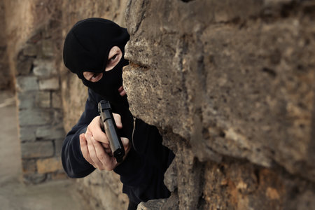 Masked man with gun hiding behind stone wall outdoors. criminal offenseの写真素材