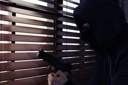 Masked man with gun spying through window blinds indoors. Dangerous criminalの写真素材