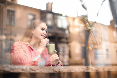 Pretty young woman with cocktail at table in cafe, view from outdoors through windowの写真素材