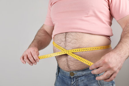 Overweight man with large belly and measuring tape on color background, closeup
